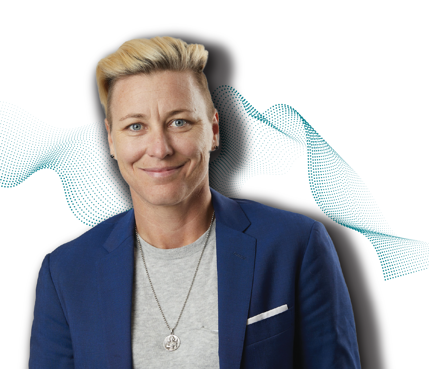 Professional headshot of Abby Wambach with short blonde hair styled in an undercut, wearing a navy blue blazer over a light gray t-shirt and a pendant necklace. The person has blue eyes and is smiling at the camera. The background features a decorative pattern of teal dots forming a fingerprint-like design
